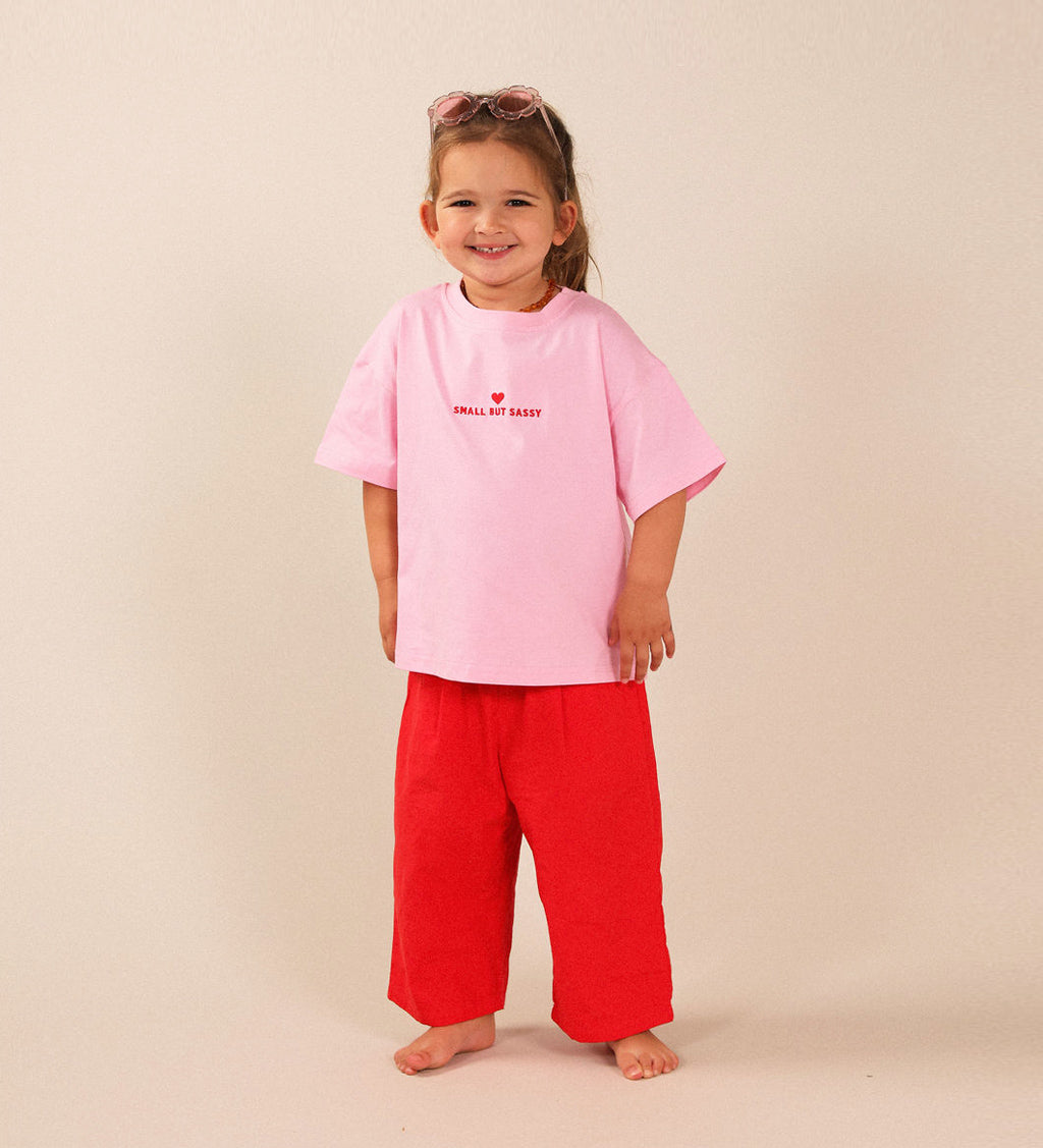 A young child standing barefoot against a plain beige studio background, wearing an oversized light pink T-shirt with SMALL BUT SASSY red text and a red heart on the chest, paired with bright red pants.