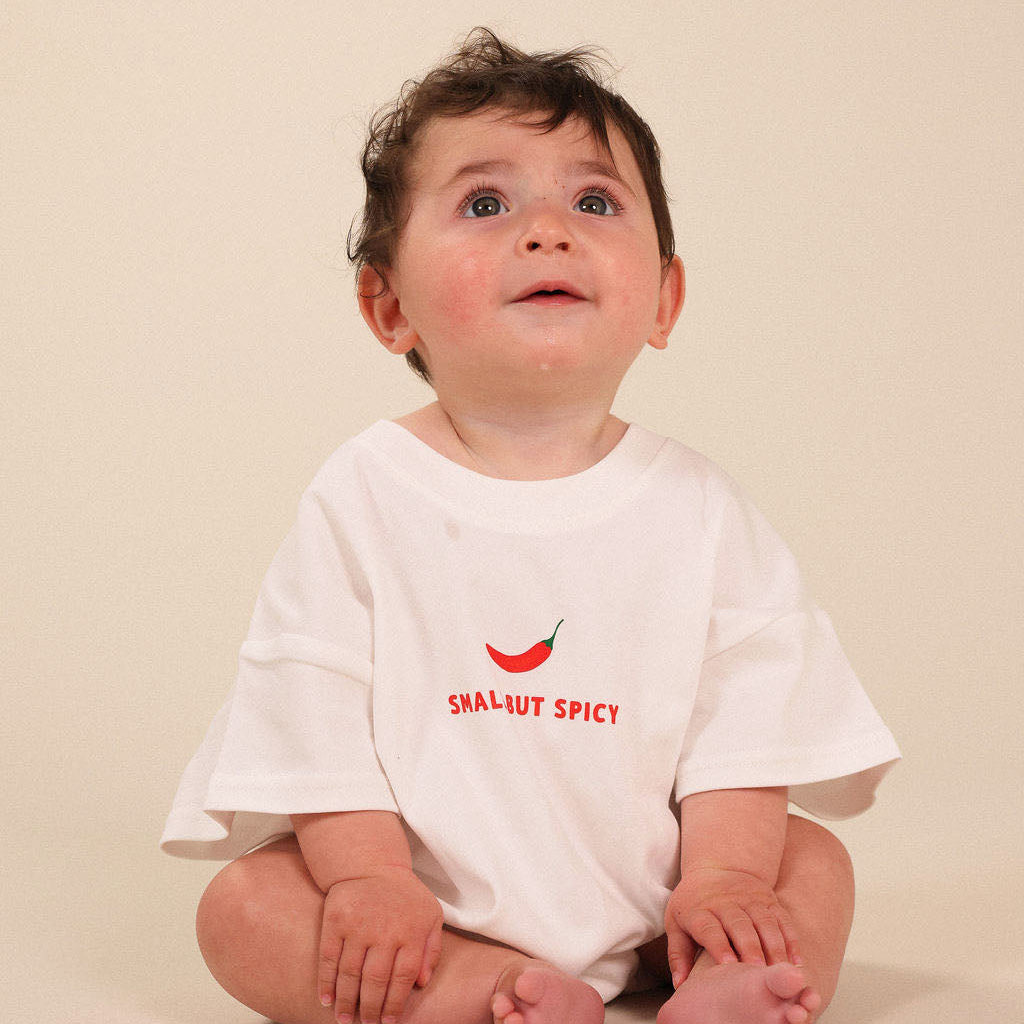 A baby sitting cross-legged wearing a white short-sleeve t-shirt with a small red chili pepper graphic and the text 'Small but SPICY' printed on the chest, set against a plain beige background.