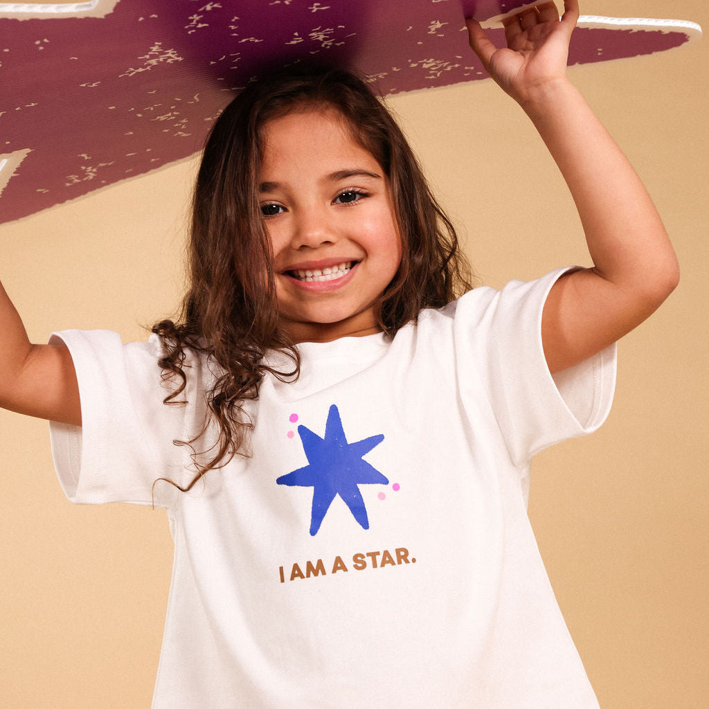 A young girl smiling and holding a purple star-shaped prop above her head, wearing a white t-shirt with a blue star graphic and the words 'I AM A STAR.' against a plain beige background.