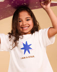 A young girl smiling and holding a purple star-shaped prop above her head, wearing a white t-shirt with a blue star graphic and the words 'I AM A STAR.' against a plain beige background.