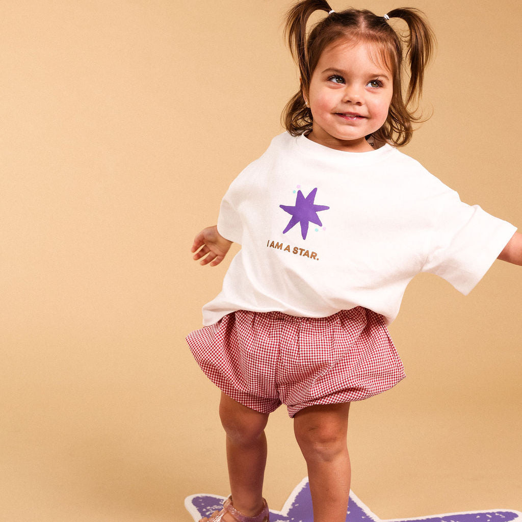 A toddler girl with pigtails stands on a purple star-shaped mat against a beige studio background. She is wearing a white graphic T-shirt with a purple star and the words 'I AM A STAR.