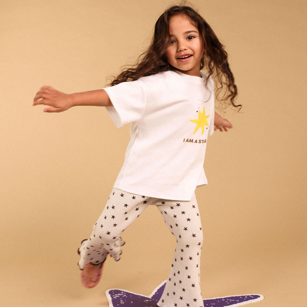 A young girl with long brown hair is captured mid-dance, balancing on a purple star-shaped rug. She is wearing a white t-shirt with a yellow star graphic and the text 'I AM A STAR' paired with white flared pants covered in small black stars.