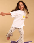 A young girl with long brown hair is captured mid-dance, balancing on a purple star-shaped rug. She is wearing a white t-shirt with a yellow star graphic and the text 'I AM A STAR' paired with white flared pants covered in small black stars.