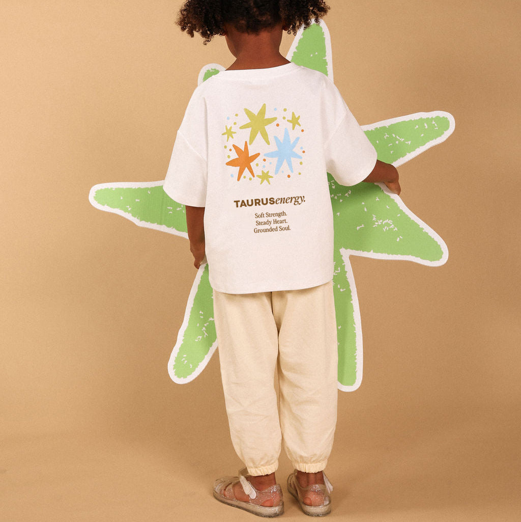 A child stands with their back to the camera wearing a white T-shirt with a colorful star graphic and 'TAURUS energy' text, paired with cream pants, against a solid beige background.