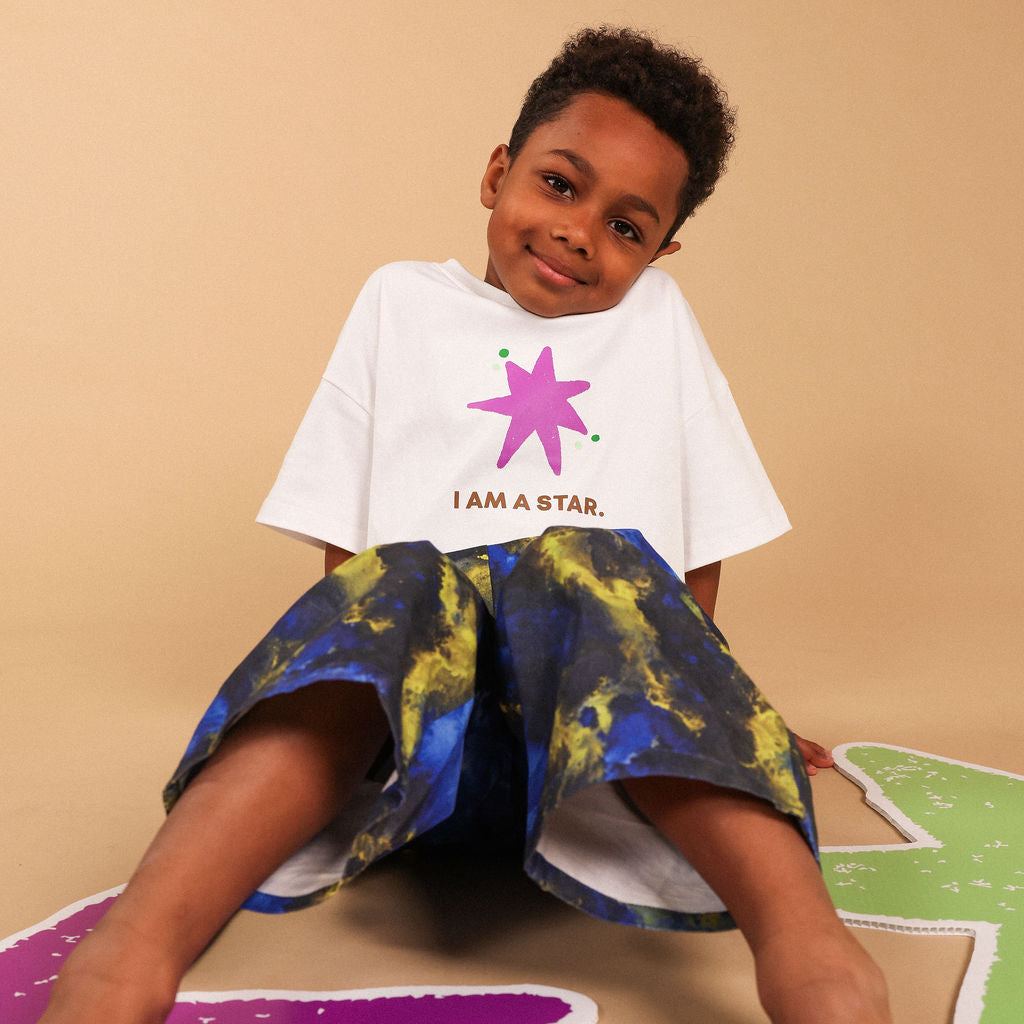 A young boy sitting on a beige floor wearing a white graphic t-shirt with a pink star and the text 'I AM A STAR.