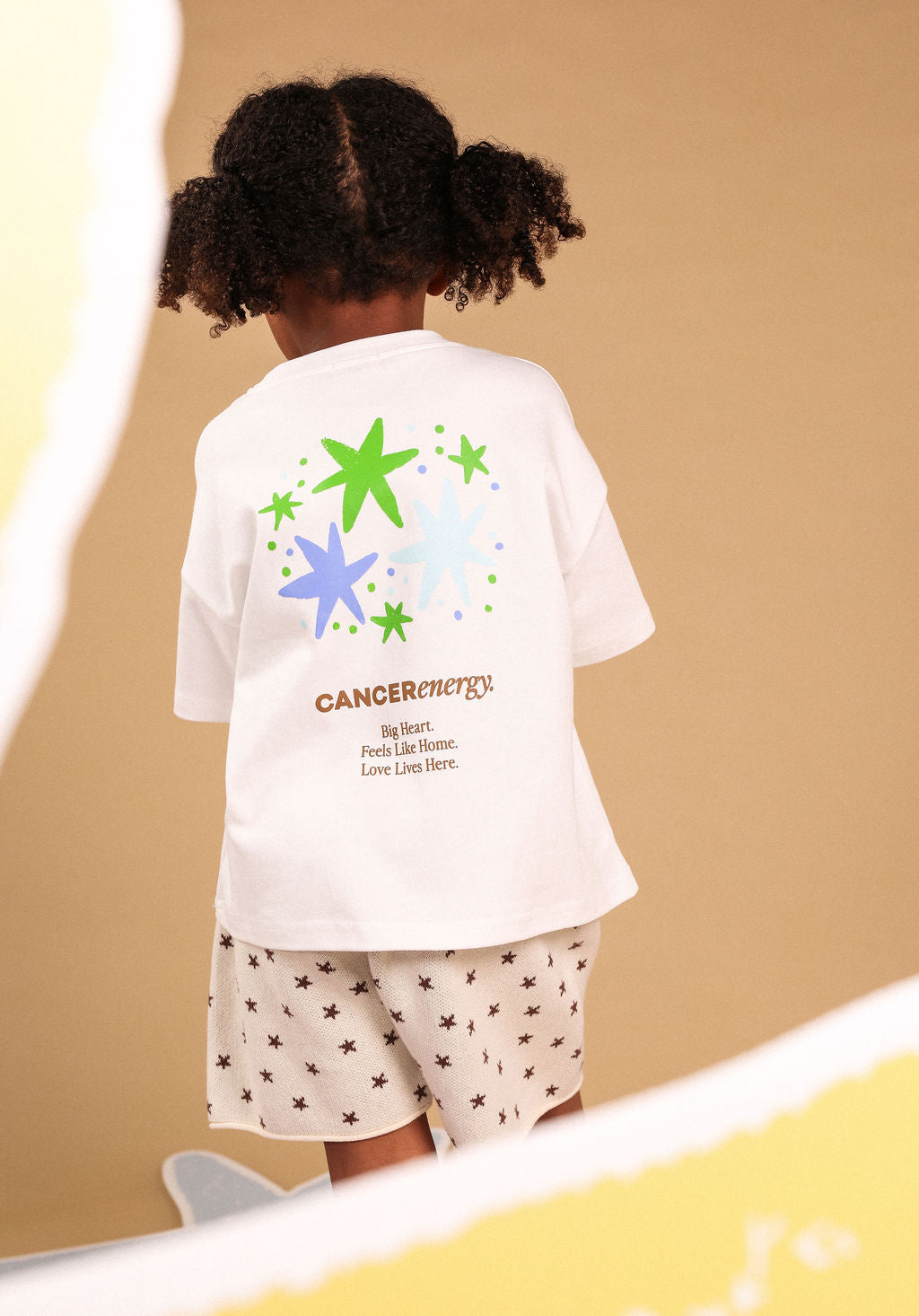 A child viewed from behind wearing a white oversized T-shirt with a colorful star graphic and the text 'CANCERenergy' on the back, paired with white shorts patterned with small dark stars, against a warm beige background.
