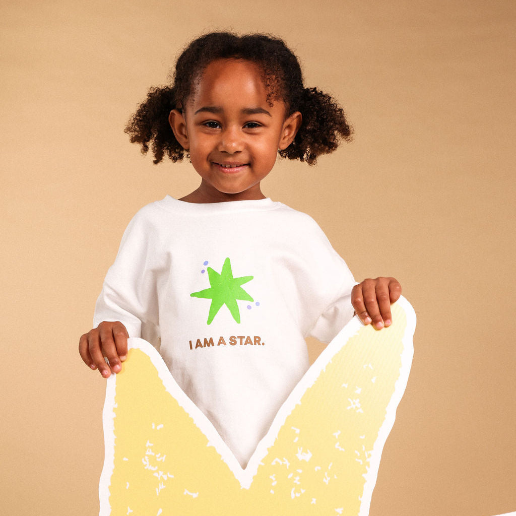 A young child with curly hair wearing a white t-shirt with a green star graphic and the text 'I AM A STAR.' holds a large yellow star-shaped prop against a solid beige background.