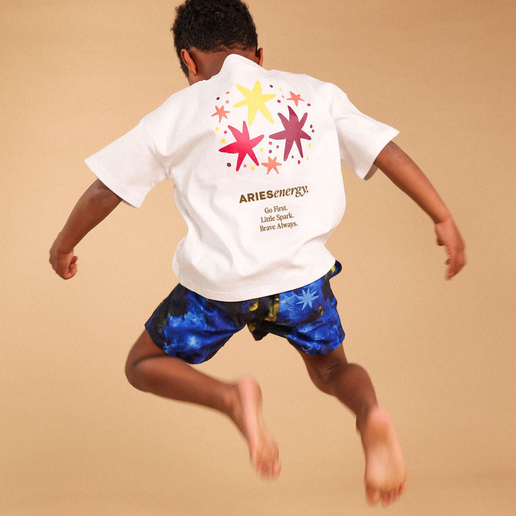 A child jumps in the air wearing a white Aries  t-shirt with colorful star graphics on the back and blue patterned trunks, set against a warm beige background with a red star-shaped chalk outline on the floor.