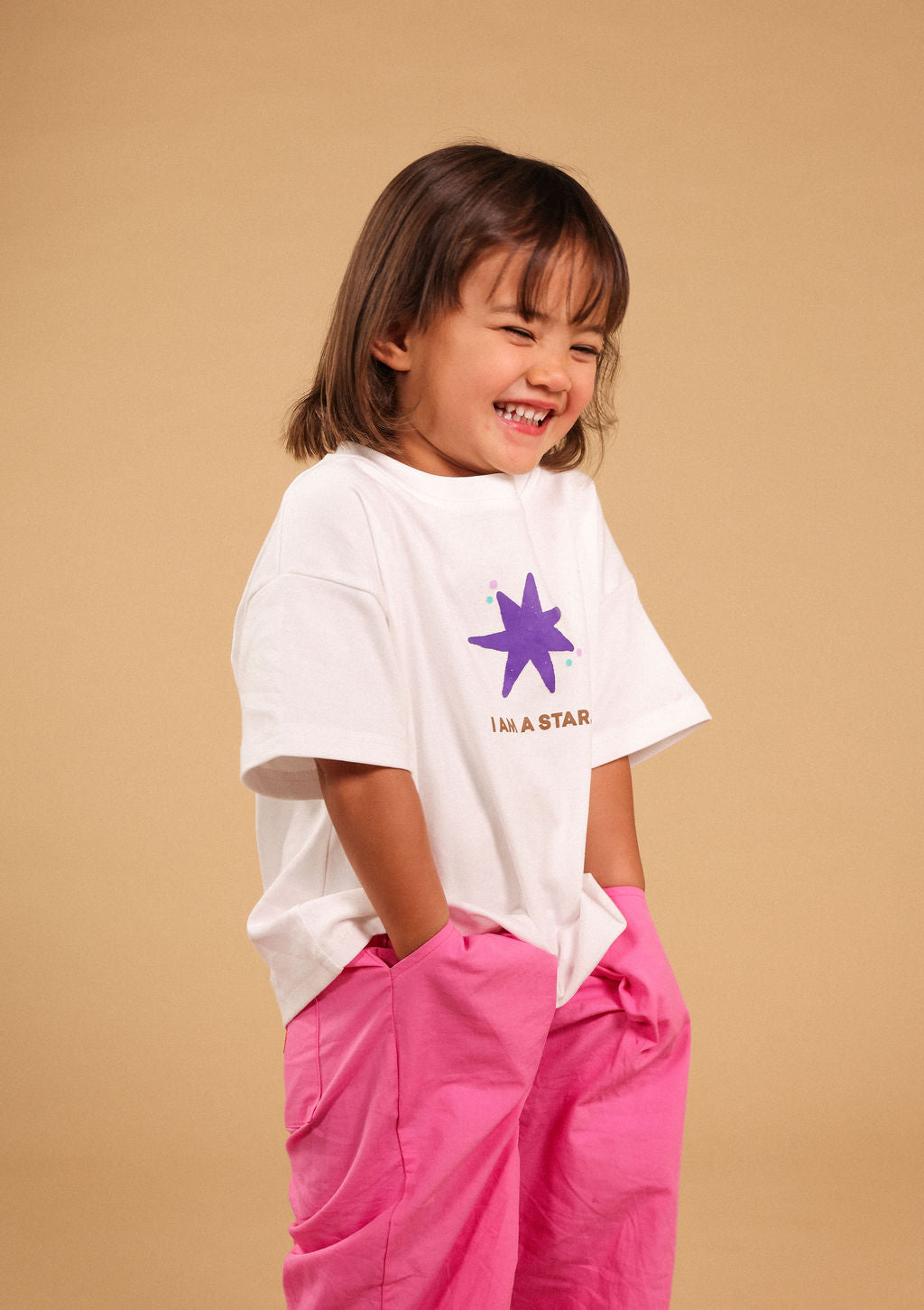 A young girl with shoulder-length brown hair smiling and laughing, wearing a white short-sleeve t-shirt with a purple star graphic and the text 'I'M A STAR' in pink pants, posed against a solid beige studio background.