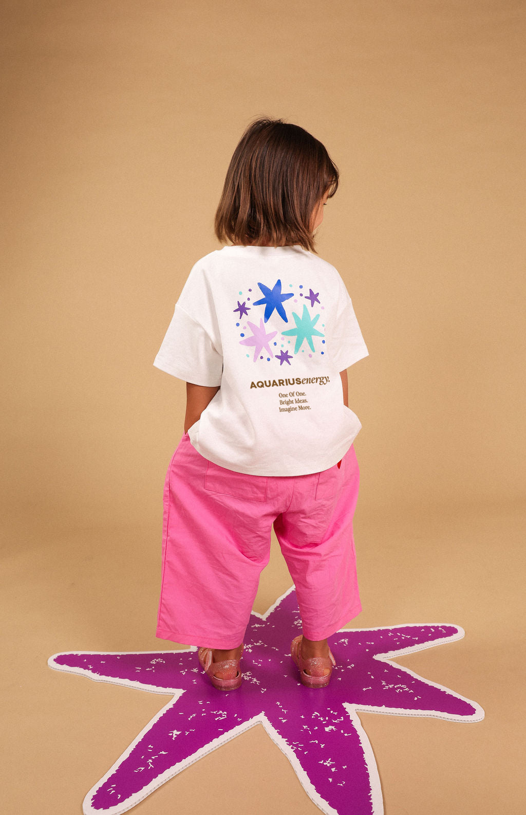 A child stands with their back to the camera wearing a white graphic t-shirt and bright pink pants, standing on a purple star shape against a solid tan background.