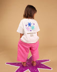 A child stands with their back to the camera wearing a white graphic t-shirt and bright pink pants, standing on a purple star shape against a solid tan background.