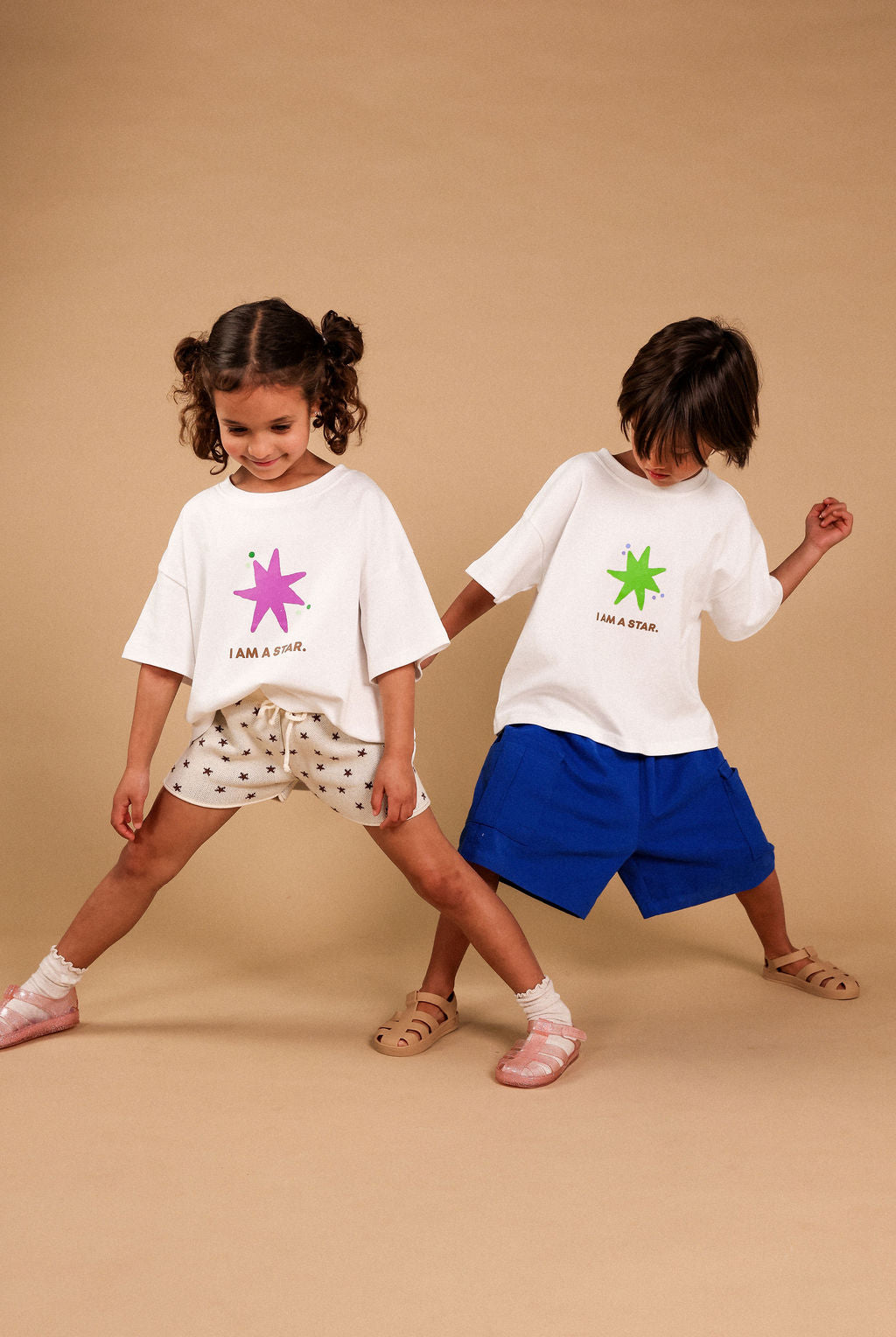 Two young children modeling matching white graphic t-shirts and shorts against a solid beige studio backdrop. The girl on the left wears a white t-shirt with a pink star graphic and the text 'I AM A STAR.