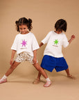 Two young children modeling matching white graphic t-shirts and shorts against a solid beige studio backdrop. The girl on the left wears a white t-shirt with a pink star graphic and the text 'I AM A STAR.