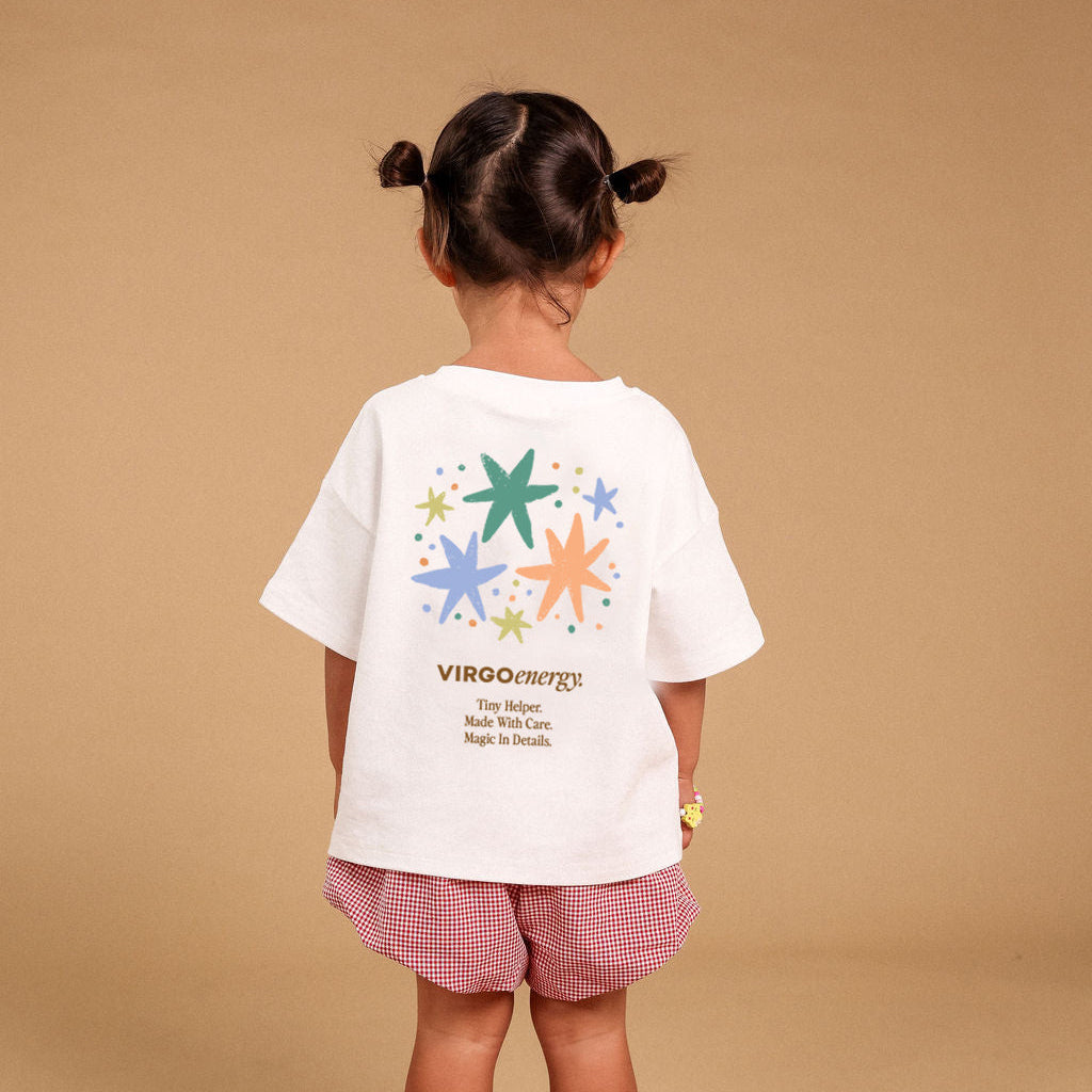 A young girl stands with her back to the camera wearing a white graphic T-shirt and pink gingham shorts, standing on a red star-shaped rug against a solid beige background.