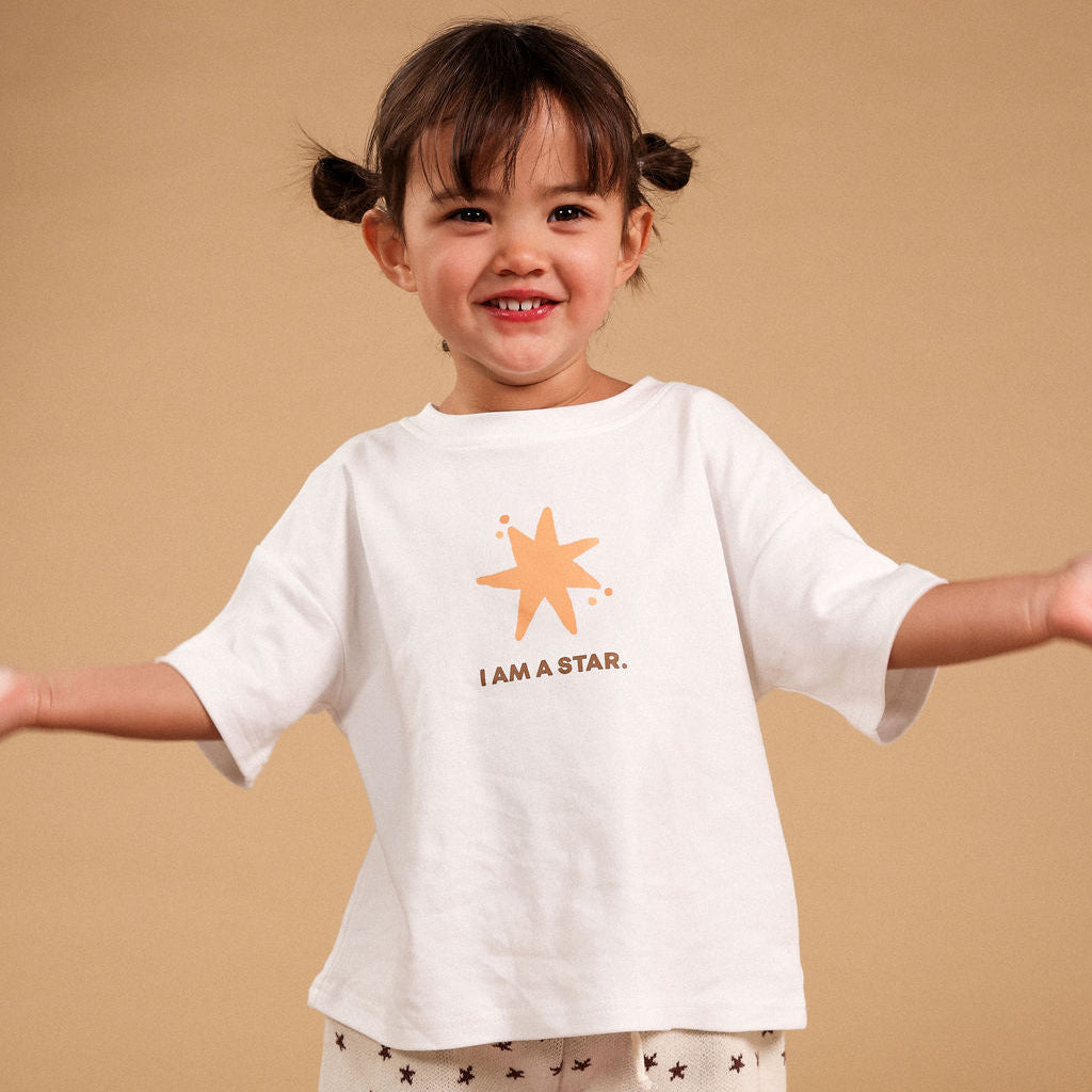 A young child with dark hair in two buns stands against a solid beige background, wearing a white short-sleeve t-shirt with a gold star graphic and the text 'I AM A STAR.' paired with cream shorts patterned with small stars.
