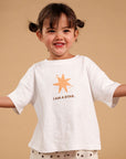 A young child with dark hair in two buns stands against a solid beige background, wearing a white short-sleeve t-shirt with a gold star graphic and the text 'I AM A STAR.' paired with cream shorts patterned with small stars.