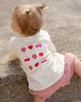 Child wearing a white shirt with red and pink heart patterns, sitting on a concrete surface.