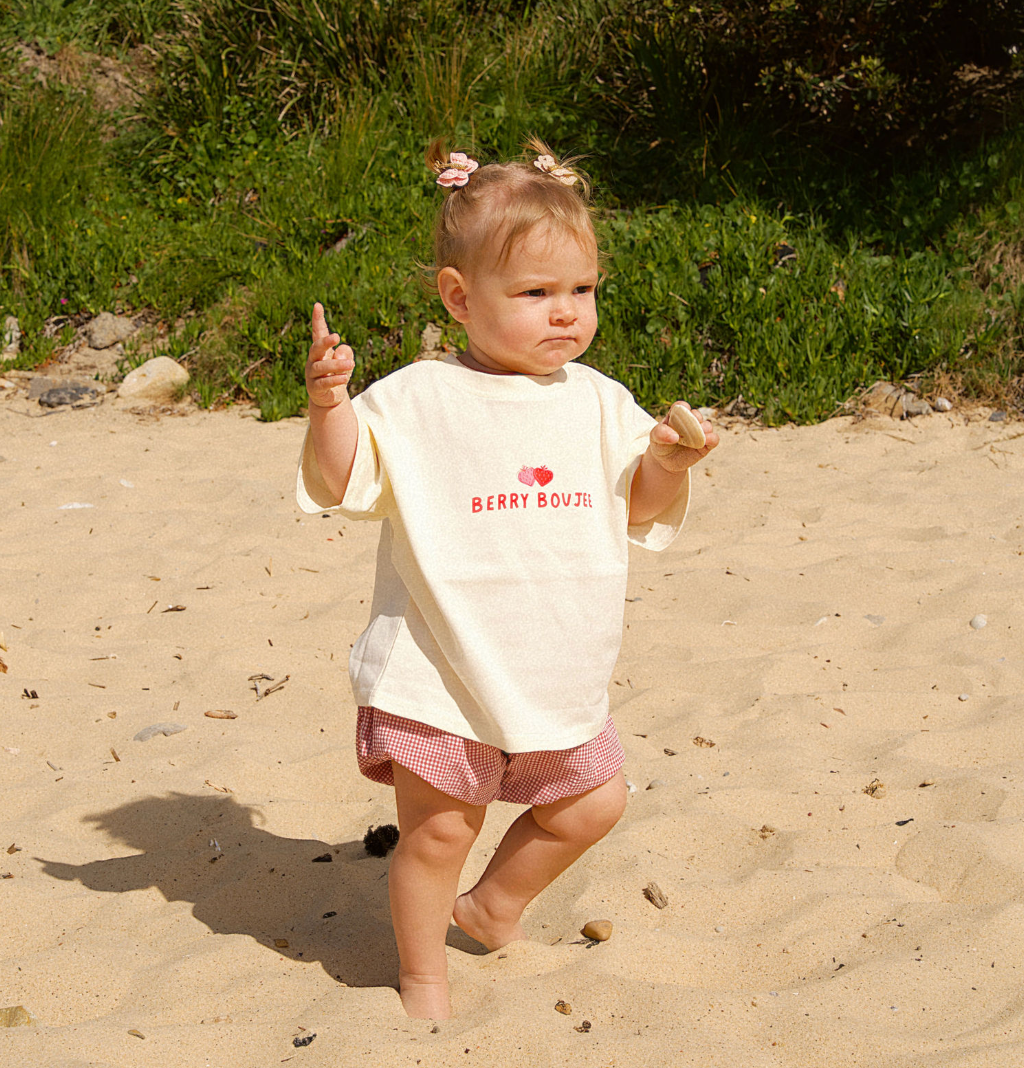 Child in a white shirt with text and pink shorts standing on sandy ground with greenery in the background