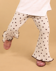 A child's lower body wearing cream flared pants with a black star pattern, a white t-shirt, and pink glittery shoes, captured mid-step against a solid beige background.