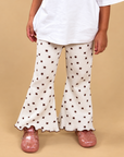 A child wearing cream flared pants with a brown star pattern, a white t-shirt, and pink clogs, standing against a solid beige background.
