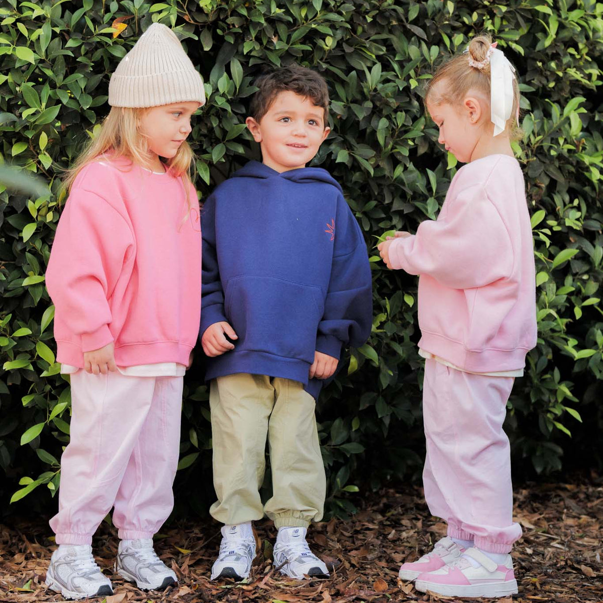 Three children in matching pink and blue outfits standing in front of a green bush.