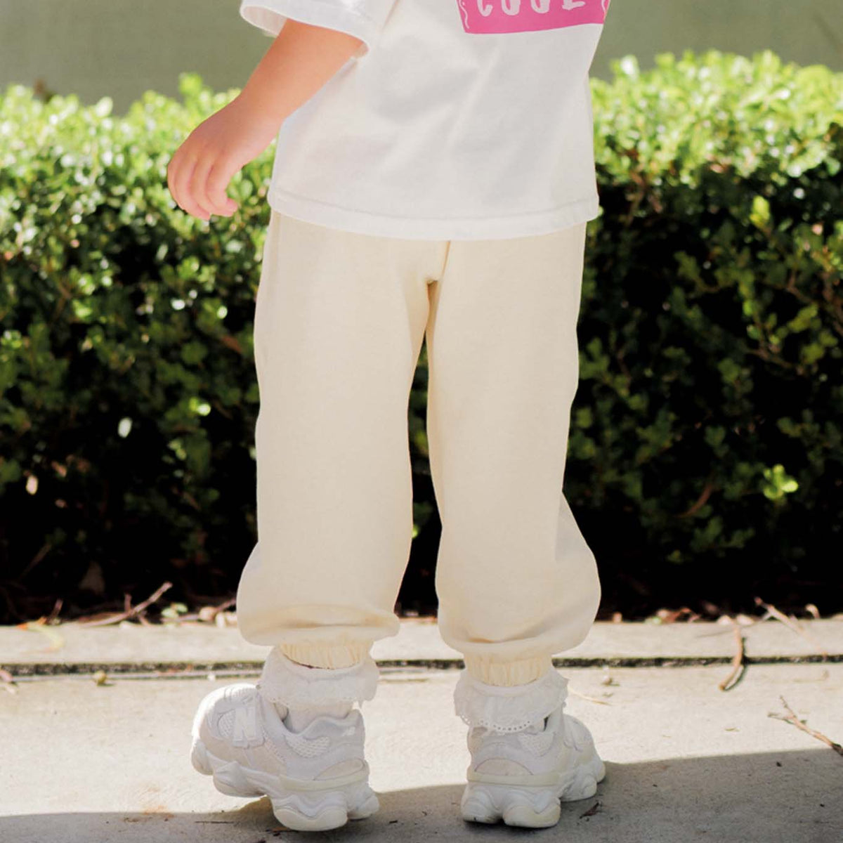 Person wearing a white shirt with pink text, beige pants, and white shoes standing on a sidewalk with green bushes in the background.