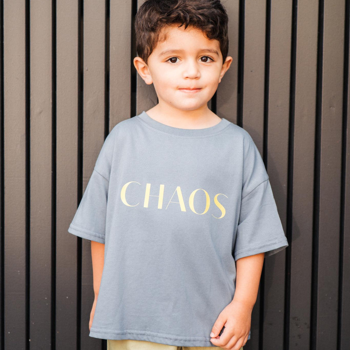 Child wearing a gray t-shirt with 'CHAOS' in yellow letters against a striped background