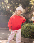 Child wearing a red sweater and pink pants standing outdoors with trees in the background