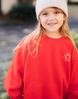 Young girl wearing a red sweatshirt with a logo and a white beanie outdoors.