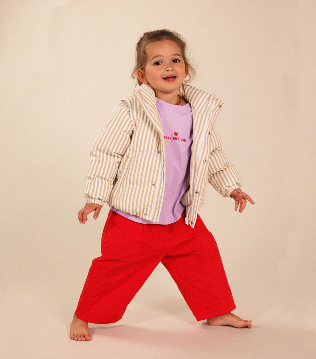 A young child modeling a light beige and white vertically striped puffer jacket over a lavender t-shirt and bright red pants, standing barefoot against a solid beige studio background.