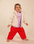A young child modeling a light beige and white vertically striped puffer jacket over a lavender t-shirt and bright red pants, standing barefoot against a solid beige studio background.