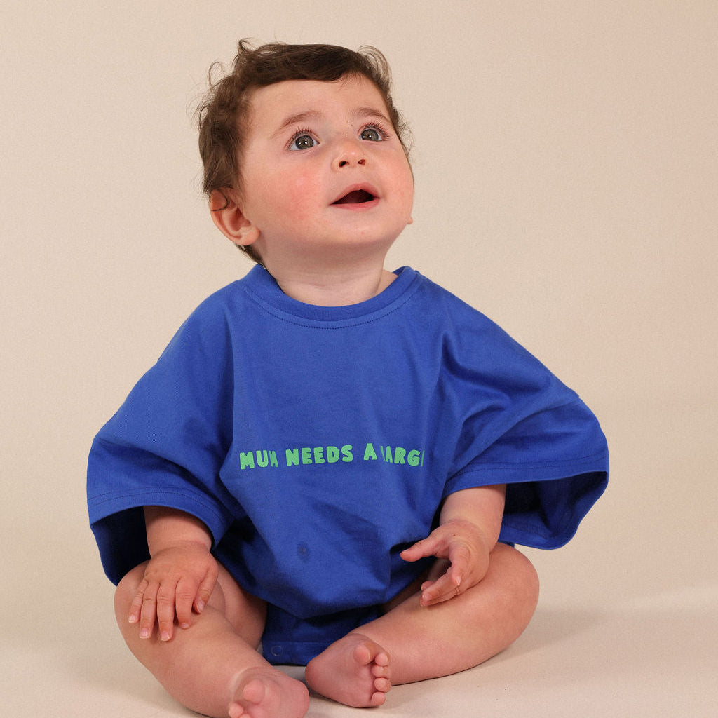 A baby sitting cross-legged wearing a bright blue oversized t-shirt with green text that reads 'MUM NEEDS A LARGER'.