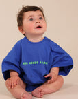 A baby sitting cross-legged wearing a bright blue oversized t-shirt with green text that reads 'MUM NEEDS A LARGER'.