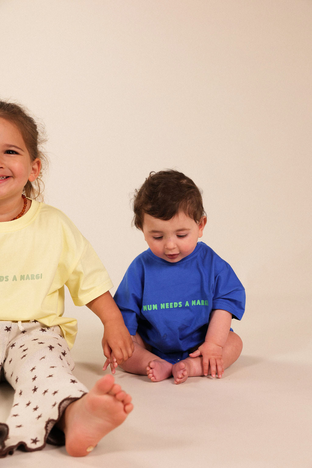 Two children sitting on a warm off-white studio floor. The toddler in the center wears a bright blue t-shirt with green text reading 'MUM NEEDS A MARGI' and white pants.