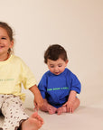 Two children sitting on a warm off-white studio floor. The toddler in the center wears a bright blue t-shirt with green text reading 'MUM NEEDS A MARGI' and white pants.