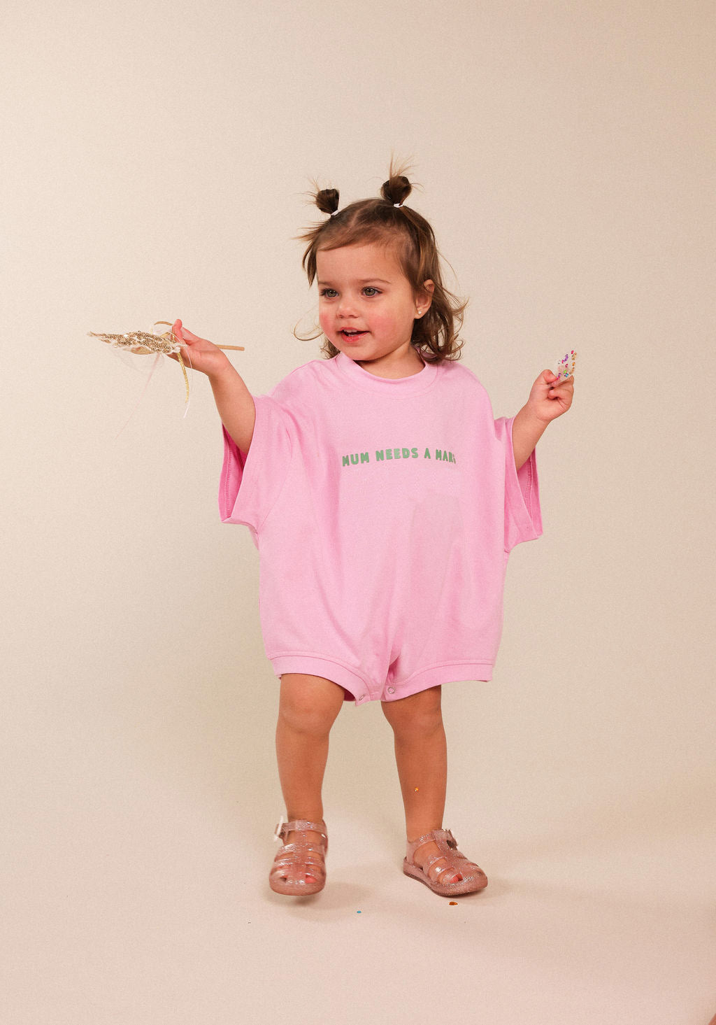 A toddler girl with two buns in her hair stands against a warm beige studio background, wearing an oversized light pink short-sleeve romper with green text reading 'MUM NEEDS A MARGI' across the chest. She holds dried grass in one hand and a small object in the other, wearing pink sandals.