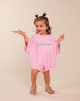 A toddler girl with two buns in her hair stands against a warm beige studio background, wearing an oversized light pink short-sleeve romper with green text reading 'MUM NEEDS A MARGI' across the chest. She holds dried grass in one hand and a small object in the other, wearing pink sandals.