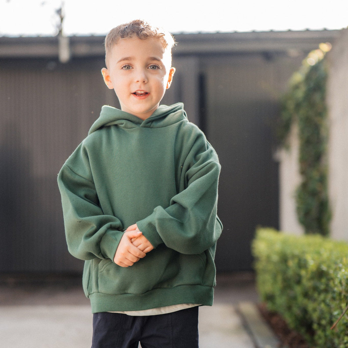 Child wearing a green hoodie and dark pants standing on a sidewalk.