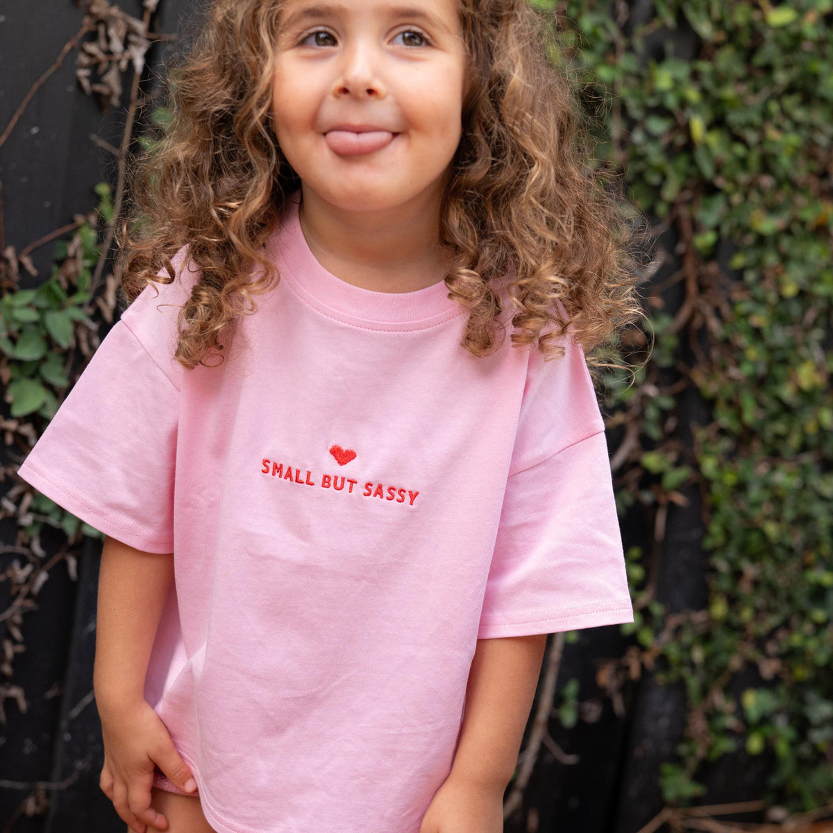 A child with curly hair is sticking out their tongue while wearing a pink t-shirt with 'SMALL BUT SASSY' embroidered in red on the front.