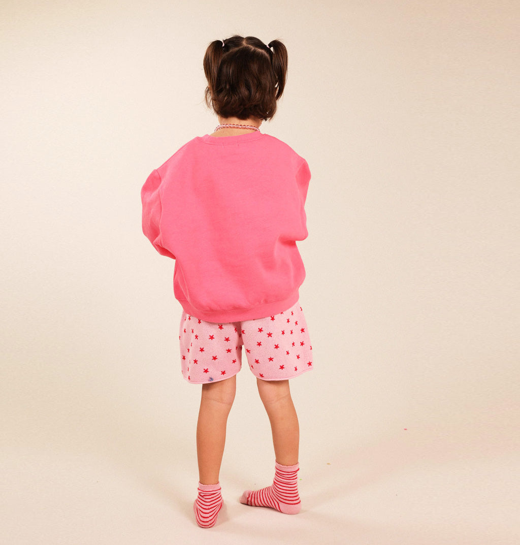 A young child viewed from behind, wearing a bright pink oversized sweatshirt, pink shorts with small red heart patterns, and pink-and-white striped socks, standing against a plain beige studio background.